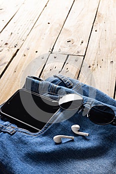 Vertical image of blue jeans with smartphone, earphones and glasses lying on wooden surface