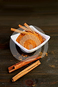 Vertical image of bowl of ground cinnamon powder with cinnamon stick on black wooden background