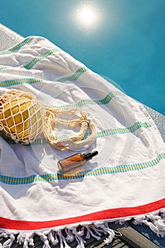 Vertical image of a brown glass jar of natural facial serum lying on a towel with a bag of fruit by the pool. Concept of