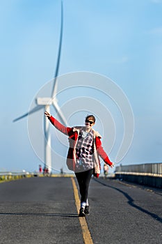 Vertical image of Caucasian woman enjoy to walk on single solid yellow line of the road on the mountain with big windmill or wind