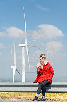 Vertical image of Caucasian woman with red coat sit on barrier of roadside and use mobile phone on mountain near windmill or wind