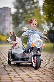 Vertical image of cute little girl  driving electrical motorcycle toy with sidecar and her dog in it, smiling