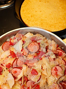 Vertical image of fried cabbage, sausage, onions, tomatoes and pan of cornbread