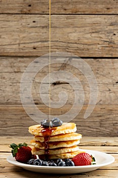 Vertical image of maple syrup pouring onto pancakes and blueberries, on wood with copy space