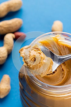 Vertical image of spoon and jar with peanut butter and nuts on blue surface