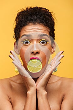 Vertical image of surprised afro american woman bulging eyes while putting half of fresh lime in mouth isolated, over yellow back