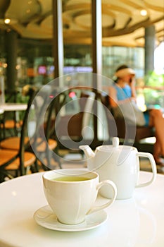 Vertical image of a white cup and teapot of green tea served on cafe`s white table