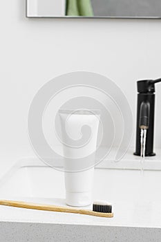 Vertical image of a white mockup of a tube of toothpaste and a toothbrush standing on a sink on the background of a stream of