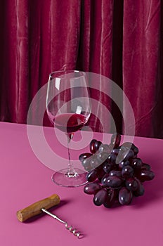 Vertical image of wineglass of red wine, corckscrew and grape on the dark pink table against folds of curtain