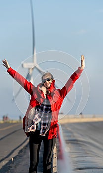 Vertical image of woman with red coat stand with rest her hands up to action of happiness in front of wind turbine or windmill in