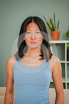 Vertical individual portrait of serious asian young woman looking at camera standing at home linving room. One photo