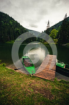 Vertical landscape photo of beautiful lake with wooden mole and green boats on sunset. Dramatic and cloudy sky over the river