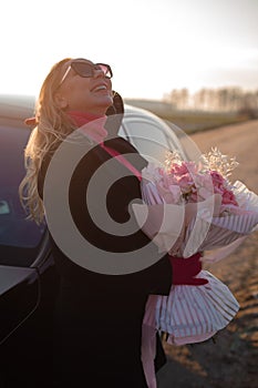 Vertical laughing fashionable blond woman in sunglasses holding posh pink rose flower decorative bouquet near black car