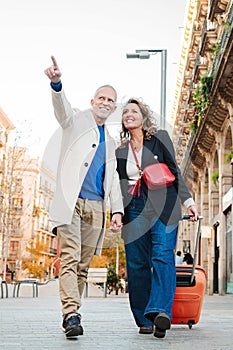Vertical. Mature tourists pointing and walking with a suitcase on vacations. Middle aged couple having fun sightseeing