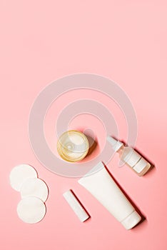 Vertical minimalistic layout with hand cream in a white tube, essential oil, lip balm, cotton pads, and a candle on a pastel pink