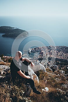 Vertical moody shot of a bald man sitting on the Srd mountain above the city of Dubrovnik, observing the area on a cold autumn day