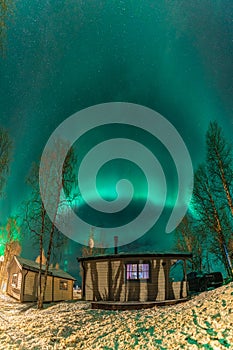 Vertical night photo of beautiful Northern lights above small wooden mountains cottage in Northern Sweden, Lapland, Joesjo. Green