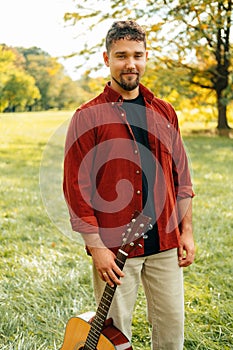 Vertical outdoors shot of a handsome man holding an acoustic guitar in park.