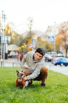 Vertical photo of an attractive man sitting on a green lawn with his cute small dog - yorkie. Blurred background, busy city street