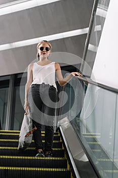 Vertical photo of blonde girl with sunglasses and a grey bag in her hand walking down an escalator