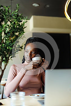 Vertical photo of calm, contemplating African American businesswoman with laptop, drink coffee or tea in internet cafe