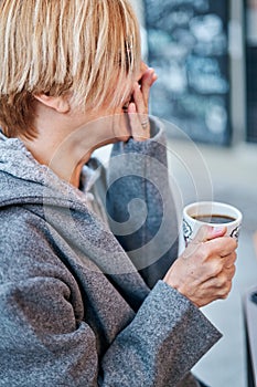 Vertical photo of Caucasian blonde woman wearing sunglasses sitting in a bar terrace with coat having a cup of coffee