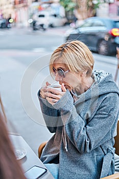 Vertical photo of Caucasian blonde woman wearing sunglasses sitting in a bar terrace with coat having a cup of coffee