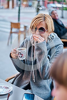 Vertical photo of Caucasian blonde woman wearing sunglasses sitting on a terrace of a bar with a coat having a cup of coffee with