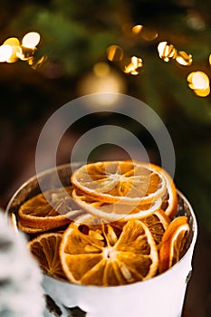 Vertical photo with a cup of dried orange slices on a dark background of a Christmas tree with bokeh from garlands