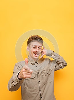 Vertical photo. Funny nerd in a shirt and glasses stands on a yellow background, shows a hand gesture of the phone and points his