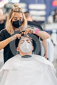 Vertical photo of a hairdresser adjusting curls of the hair of a man with mask