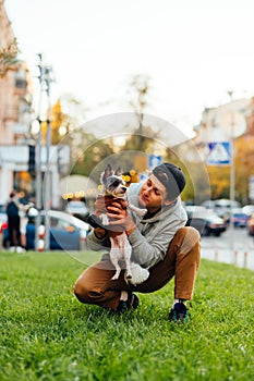 Vertical photo of a joyful funny young male dog-owner holding his cute doggy - york terrier, sitting on a green lawn near the busy
