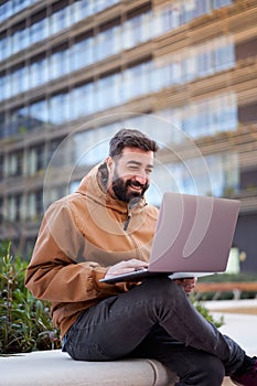 Vertical photo of a man using laptop outdoors. Businessman with a nice smile works remotely on the street.