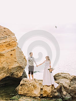 Vertical photo of the newlywed couple holding hand on the cliff at the background of the sea.