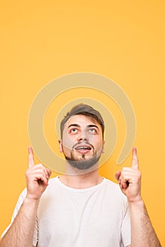 Vertical photo. Portrait of a surprised man wearing a white T-shirt, standing on a yellow background, looking up and showing his