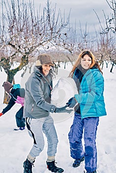 Vertical photo of two young caucasian girls wearing gloves, enjoying and carrying a snowball to make a snowman