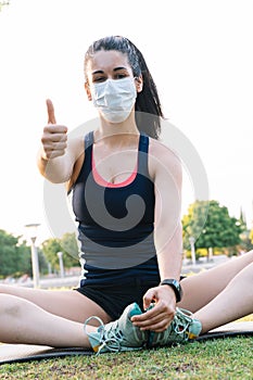 Vertical photo of a woman with mask stretching on the ground and doing a gesture to be fine outdoors