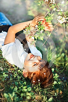 Vertical picture of 7 years old smiling girl with stylish short haircut laying down in grass under blooming cherry tree in spring