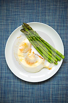 Vertical picture of baked roasted fresh green asparagus with fried eggs under parmesan cheese in big white plate on blue