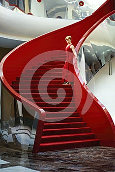Vertical picture of beautiful elegant woman with short haircut in red evening dress standing on red stairs. Happy elegant short-