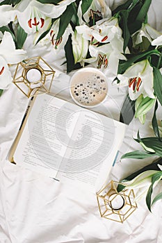 Vertical picture of opened book, coffee cup, candlestick, glasses and fresh flowers on the white bed. Flat lay, top view