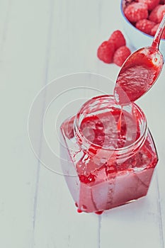 Vertical picture of raspberry jam in a jar with a spoon in it on the table near raspberries