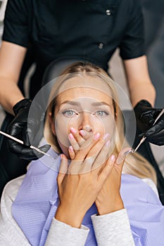Vertical portrait of afraid blonde female patient closing mouth with hands, preventing examining of teeth, because of