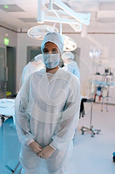 Vertical portrait of African-American female doctor in surgical mask and hat in operating room. Nurse standing posing
