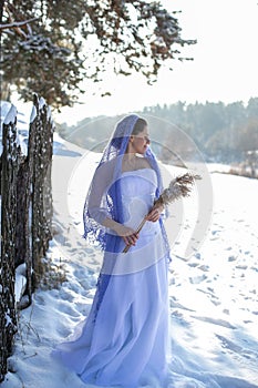 Vertical portrait against the winter landscape of a slender brunette in a long white dress and a beautiful openwork lilac shawl