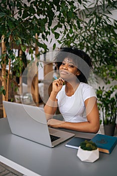 Vertical portrait of attractive African American young woman holding pen in hand, smiling looking at camera sitting at