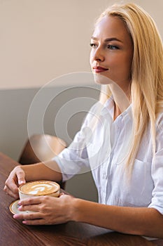 Vertical portrait of attractive young blonde woman holding cup of hot latte coffee with beautiful pattern sitting at