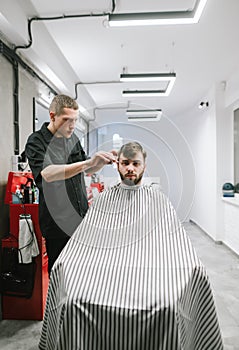 Vertical portrait of a barber and his client in a bright modern barbershop. Hairdresser cuts a bearded man`s scissors. Creating