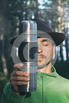 Vertical portrait of bearded millennial man in felt hat with closed eyes holding stainless thermos near his face on woods