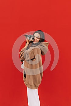 Vertical portrait of a beautiful dog-owner female in a hoodie hugging her adorable york terrier in front of the isolated red wall